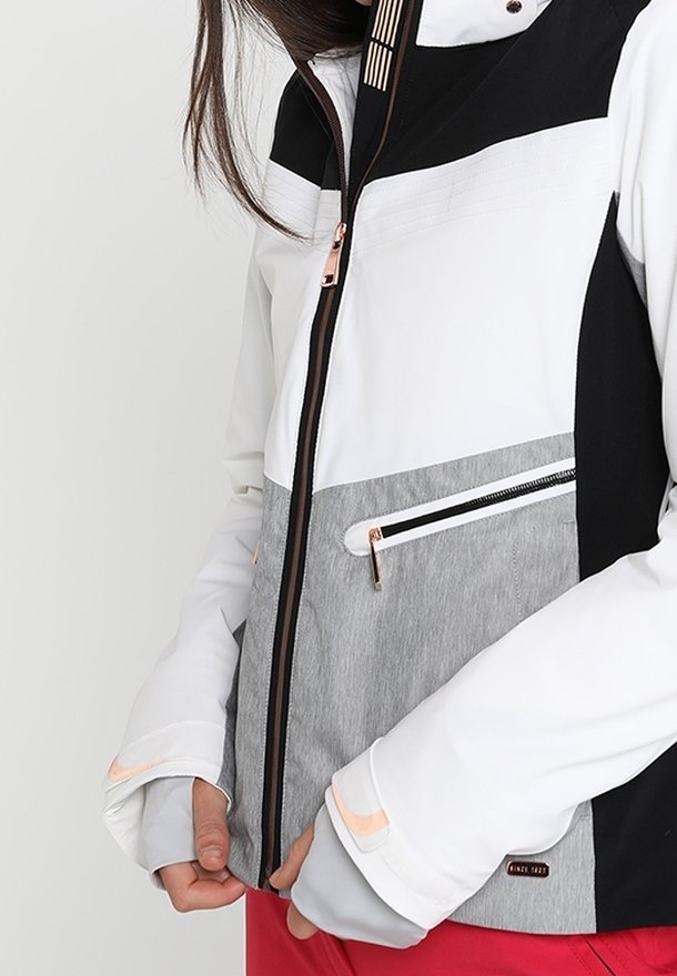 Femme portant une veste zippée blanche, grise et noire avec des trous pour les pouces, tenant l'ourlet inférieur au-dessus d'un pantalon rouge, debout devant un fond uni.