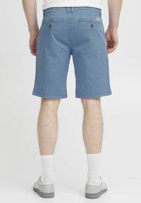 Shorts en coton bleus pour hommes, longueur genou, avec deux poches arrière à boutons et une texture lisse et uniforme. Portés avec des chaussettes blanches et des chaussures grises.