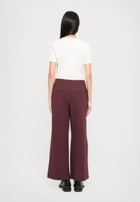 Une chemise blanche à manches courtes avec un petit logo, associée à un pantalon large bordeaux avec des poches arrière, porté avec des chaussures noires.
