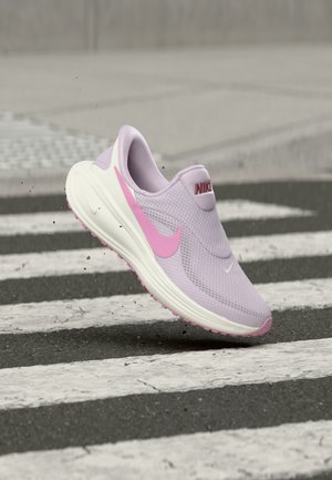 Λιλά και ροζ αθλητικό παπούτσι Nike στον αέρα πάνω από μια ασπρόμαυρη διάβαση πεζών σε έναν ασφαλτοστρωμένο δρόμο.