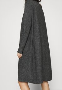 Dlouhý, oversized svetr v tmavě šedé pletené tkanině. Má žebrované manžety a boční kapsy, hladkou texturu a volný siluetu.