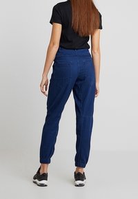 Blå joggerbyxor med elastiska muddpartier, med sid- och bakfickor, tillverkade av ett lätt material. Bärs med en svart topp och sneakers.