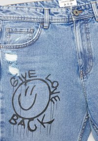 Jeans in denim leggero blu chiaro con dettagli strappati, caratterizzati da una grafica circolare nera con la scritta "DONA AMORE" e una faccina sorridente.