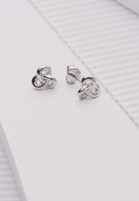 Zilveren knoop studs met een gepolijste afwerking, met een ontwerp van drie lussen en een veilige post achteraan, weergegeven op textuur papier in het wit.