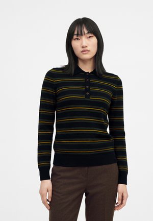 Junge Frau mit glatten schwarzen Haaren, die ein schwarz-grün gestreiftes Langarm-Poloshirt und braune Hosen vor weißem Hintergrund trägt.