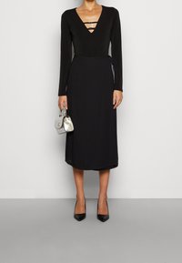 Zwarte jurk met lange mouwen en een diepe V-hals, voorzien van drie horizontale uitsparingen, ceintuurtje op de taille, gecombineerd met een kleine zilveren handtas en zwarte hakken.