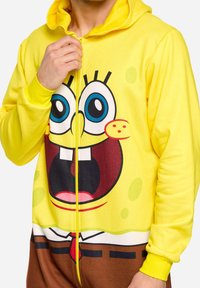 Geltona hoodie su šypsena piešinio veikėjo grafika. Minkšta medžiaga su užtrauktuku priekyje, guminiais rankogaliais ir žaismingu dizainu.