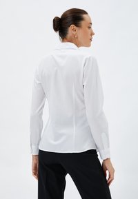 Biała koszula z długim rękawem i kołnierzykiem, o dopasowanym kroju i zapinanych mankietach, pokazana z tyłu na neutralnym tle.
