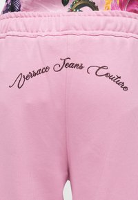 Roze trenirke imaju vezeno "Versace Jeans Couture" u tamnom, sjajnom pismu duž stražnjeg dijela. Imaju elastičnu traku u struku i mekani materijal.