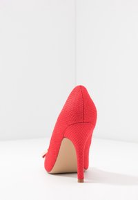 Escarpin rouge en matériau tissé texturé, à bout pointu et talon aiguille mince. Présente un petit accent sur le côté.