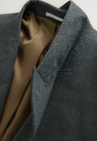 Blazer grise texturé avec une doublure en satin brun lisse, col taillé et accents brodés à la main sur le revers.