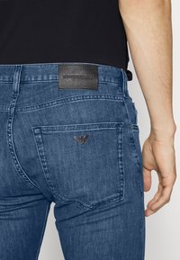 Modré džínové kalhoty s klasickým střihem, s černou koženou štítkovou páskou a kovovým logem na zadní kapse.