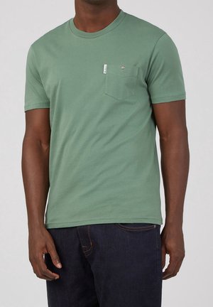 Homme portant un t-shirt vert clair à manches courtes avec une poche poitrine et un jean bleu foncé, faisant face à l'appareil photo sur un fond uni.