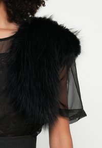 Mulher vestindo um top de renda preta translúcida com mangas curtas e um xale de pele sintética preta sobre o ombro, contra um fundo branco.