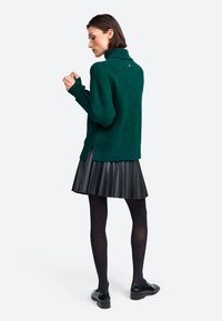 Grüner, dicker Strickpullover mit hohem Kragen, seitlichen Schlitzen, kombiniert mit einem schwarzen, plissierten Kunstlederskirt und schwarzen Strumpfhosen.