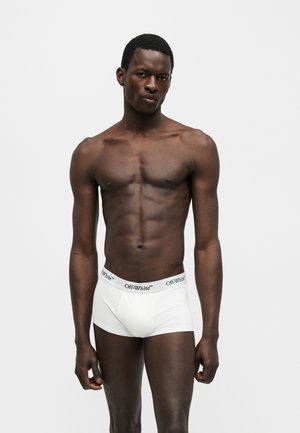 Muž bez trička s muskulatúrnou postavou, ktorý má na sebe biele boxerky značky Off-White a stojí na jednoduchom svetlom pozadí.