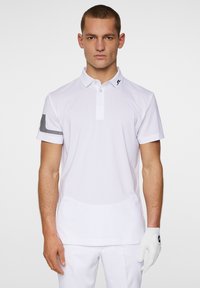 Weißes Poloshirt mit strukturierter Oberfläche, zwei Knöpfen und einem schwarzen Logo am Kragen. Der linke Ärmel hat ein schwarz-weißes gestreiftes Design.