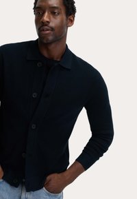 Mann in einem schwarzen, knopfengeknöpften Cardigan über einem schwarzen Hemd, mit hellblauen Jeans, die Hände in den Taschen, leicht zur Seite schauend.