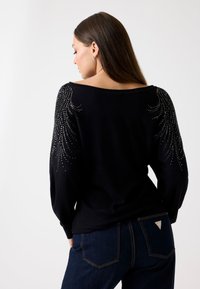 Schwarzer Pullover mit weitem Ausschnitt, verziert mit Strasssteinen in einem federartigen Design auf den Schultern. Getragen mit dunkelblauen Jeans.