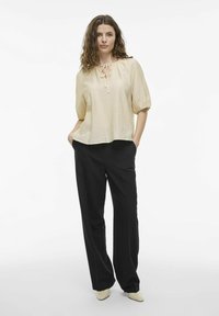 Blusa color crema con un acabado texturizado, escote redondo y detalle de lazo en la parte delantera, combinada con pantalones de pierna ancha negros.