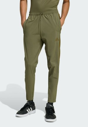 Muž v olivovo zelených zúžených nohaviciach s bočnými vreckami a čiernych teniskách Adidas s bielymi pruhmi, stojaci s rukami vo vreckách.