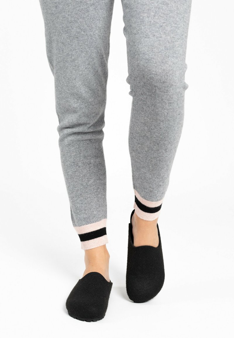 Pantofole nere in feltro sui piedi nudi, abbinate a joggers grigi con polsini a coste rosa e neri. Design semplice, facile da indossare, con punta arrotondata.