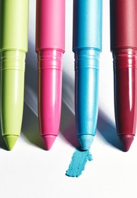 Four colorful pastel crayons—green, pink, blue, and red—lined up with the blue crayon drawing a thick line on white paper.