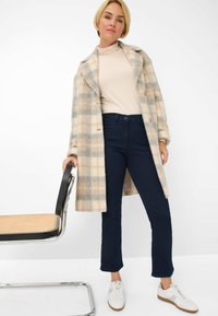 Manteau en laine à carreaux beige et gris, col roulé crème, jeans bleu foncé, et baskets blanches. Le modèle se tient à côté d'une chaise à assise en cannage.