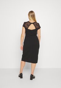 Schwarzes, figurbetontes Kleid mit Spitzenkurzärmeln und einem offenen Rückendetail. Weicher Stoff, knielange Silhouette, kombiniert mit Schuhen mit Blockabsatz.