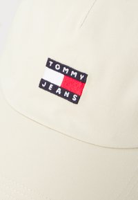 Kremfarget caps med en strukturert tekstur. Har en rektangulær brodert logo-patch i svart, hvit og rød med teksten "TOMMY JEANS".