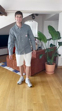 Csíkos, világoskék és fehér gombos ing, fehér rövidnadrággal párosítva. Szürke sportcipő. Belső tér, egy növénnyel és kanapéval.