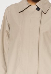Beige trenchcoat med struktureret krave, enkelt sort knap og lodrette sømdetaljer foran. Glat tekstur, letvægtsmateriale.