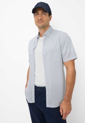 Mann trägt eine dunkelblaue Kappe, ein hellblaues kurzärmeliges Hemd über einem weißen T-Shirt und dunkelblaue Hose, steht mit den Händen in den Taschen.