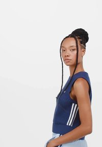 Sininen neulottu ilman hihoja oleva toppi, jossa on tekstuuri, valkoiset sivuraidat ja tiukka helma. Malli esittää neutraalia ilmaisua.