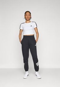 T-shirt bianco cropped con strisce nere sulle spalle e un piccolo logo, abbinato a pantaloni della tuta grigio scuro con strisce bianche di accento.