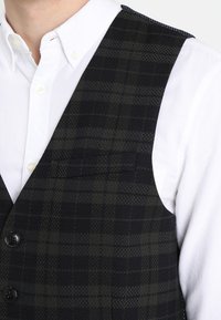 Ciemna kamizelka w kratę z tkaniny tkanej o teksturowej powierzchni. Posiada sześć guzików i kieszeń na piersi. Noszona na białej koszuli.