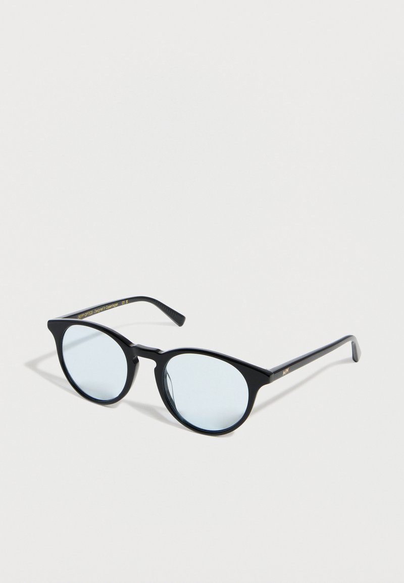 Lunettes rondes noires avec montures fines et verres teintés bleu clair posées sur une surface blanche.