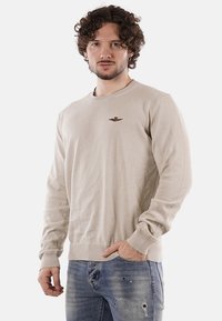 Uomo con capelli ricci che indossa un maglione beige con un piccolo emblema ricamato e jeans blu strappati, in piedi di fronte a uno sfondo bianco.