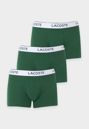 Hombre de pie descalzo, con unos bóxeres verdes con cinturilla blanca etiquetada como "Lacoste,", mirando hacia un lado sobre un fondo liso.