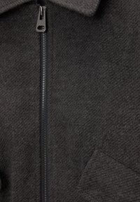 Veste grise en tissu texturé semblable à de la laine, avec une fermeture éclair apparente et une poche à rabat sur le côté gauche.