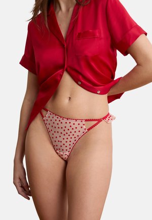 Woman wearing red satin short-sleeve shirt and sheer nude panties with red heart patterns and side cutout details, standing against white background.