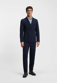 Marineblauw colbert met dubbele rij knopen en twee voorzakken, daaronder een lichtblauw textuurhemd, en zwarte loafers. Een nauwsluitende pasvorm en strakke lijnen.