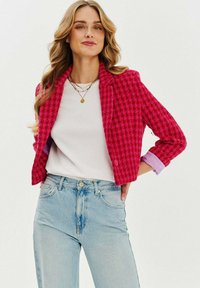 Ružový houndstooth blejzer so štruktúrovaným strihom, dvoma gombíkmi a detailmi prehnutých manžiet, nosený cez biely top, skombinovaný so svetlomodrými džínsami.