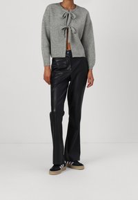 Cardigan grigio lavorato a maglia con due fiocchi sul davanti, abbinato a pantaloni in pelle neri a vita alta e sneakers nere con strisce bianche.