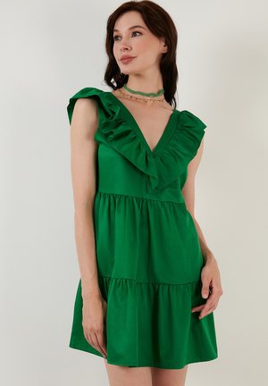 Green short dress with ruffled shoulder straps, deep V-neckline, and tiered skirt. Smooth fabric texture and playful silhouette.