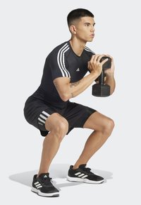 Zwarte sportshirt met witte strepen, zwarte shorts, zwarte dumbbell en zwarte sneakers met witte accenten. Persoon voert een squat uit.