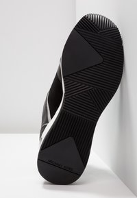 Semelle de sneaker noire avec des motifs de grip texturés et la marque "MICHAEL KORS" sur le talon, inclinée contre un fond blanc.