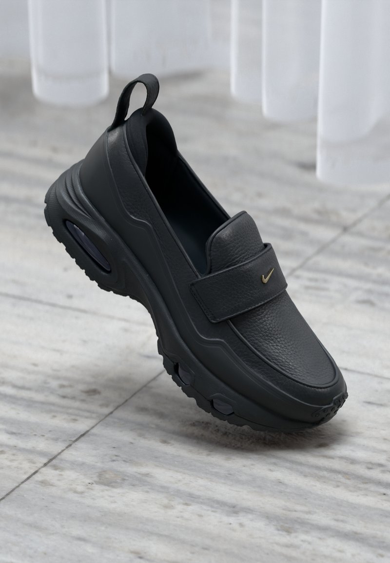 Chaussure à enfiler en cuir noir avec une semelle plate, une finition texturée, une partie supérieure rembourrée et un accent de logo doré. Conçue pour le confort et la facilité d'utilisation.