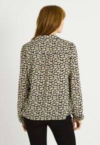 Blusa de manga larga en tela negra translúcida con patrón floral en crema, con un corte recto y detalles de botones en la parte trasera.