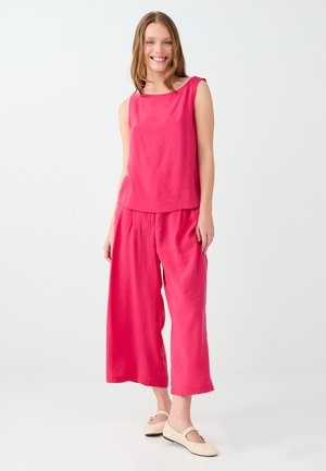 Femme souriante aux longs cheveux portant un haut rose sans manches et un pantalon large rose, debout dans des chaussures plates couleur crème.